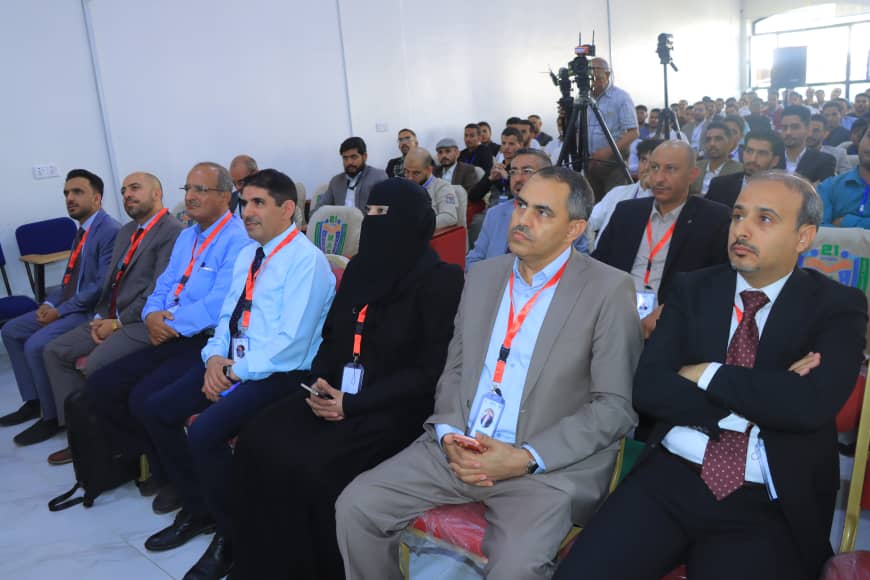 Audience seated in a conference hall, listening to a presentation; includes men in suits, a woman in a black niqab, and event cameras in the background.