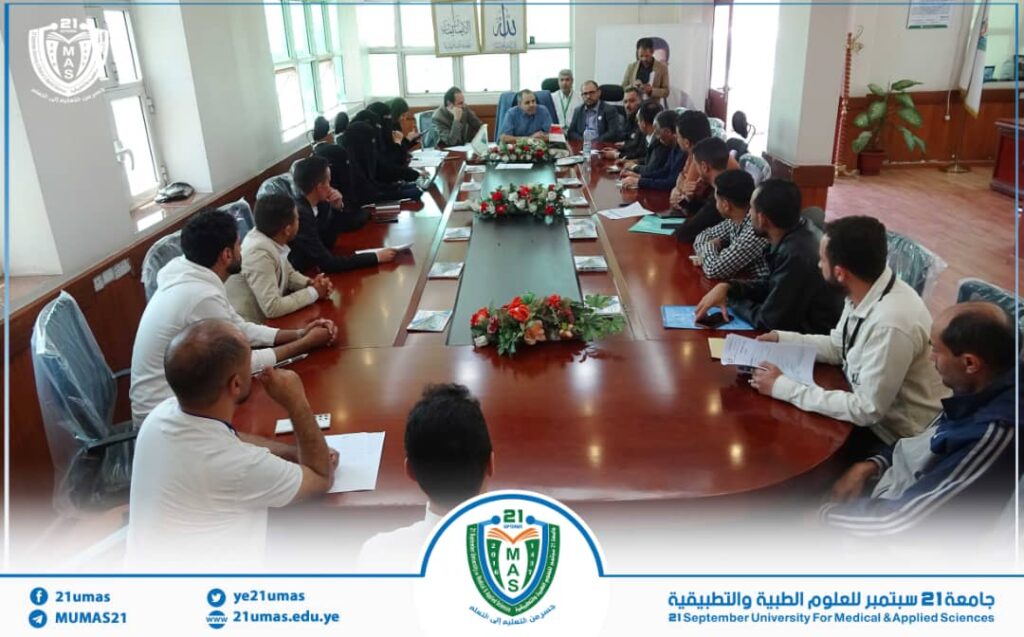 Large conference room meeting with about 25 people around a long table, a presenter at the far end leads the discussion; banners and a university emblem are visible in the scene.