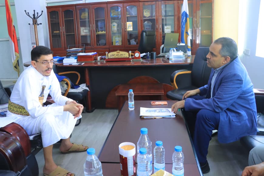 Two men sit across a conference table in an office, having a discussion; one in a white traditional outfit, the other in a blue suit. Bottled water and papers on the table.