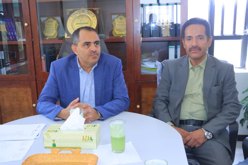 Two men in suits sit at a round table in an office, with a tissue box and a green beverage on the table and a cabinet with plaques behind them.