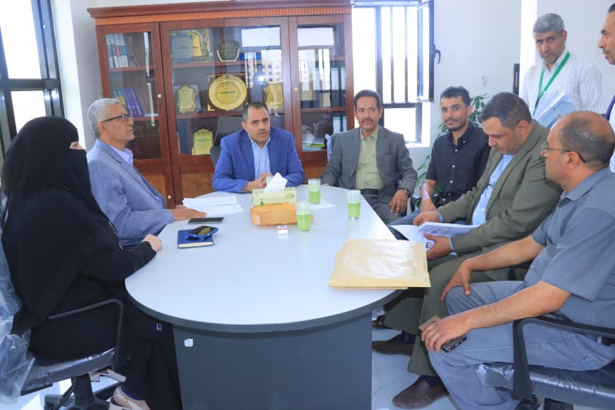 A diverse group of eight people sits around a round conference table in an office, discussing documents and a tissue box at center.