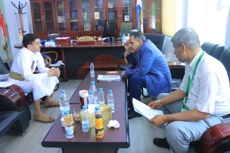 Three men sit around a coffee table in an office, discussing with papers and bottles on the table.