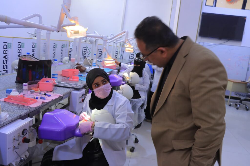 Dental students in white coats and masks working at a clinic bench with dental tools and chairs, while a man in a brown blazer observes nearby.