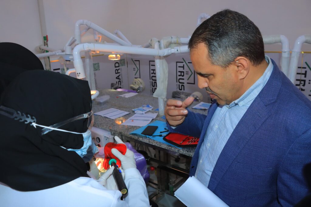 Medical professional in gloves handles dental instruments near a patient in a clinic setting with white pipe framework in the background.