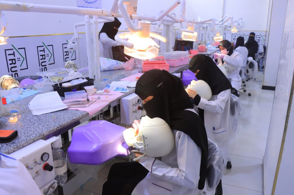 Dental students or clinicians in white coats and hijabs working at a busy dental clinic, illuminated by overhead dental lights and equipped with multiple workstations and tools.