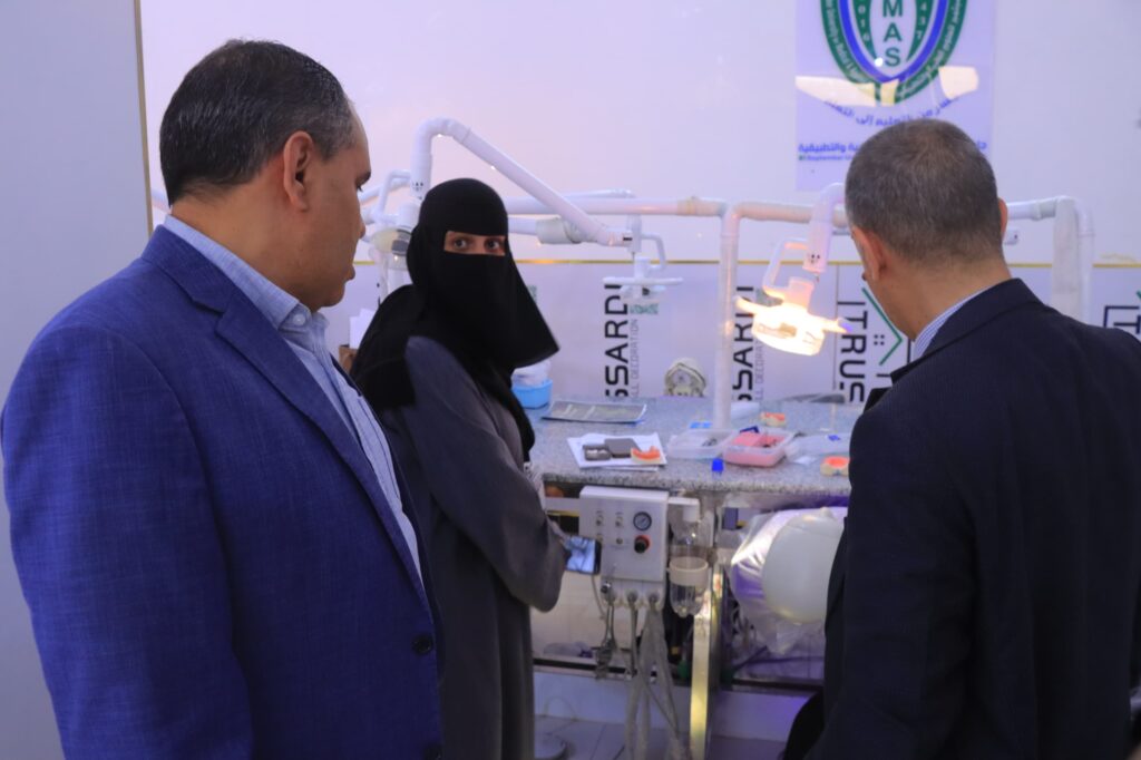 Three adults in suits stand near a dental workstation with hoses, lights, and trays in a clinic.