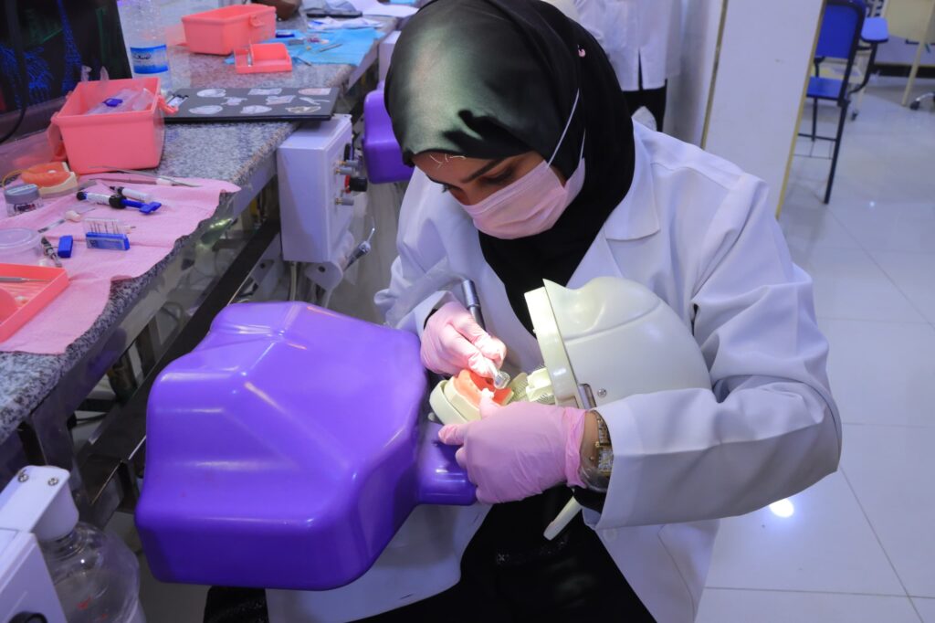 Female dental student in a white coat and pink mask works on a dental mannequin at a clinic station with tools and pink trays nearby.