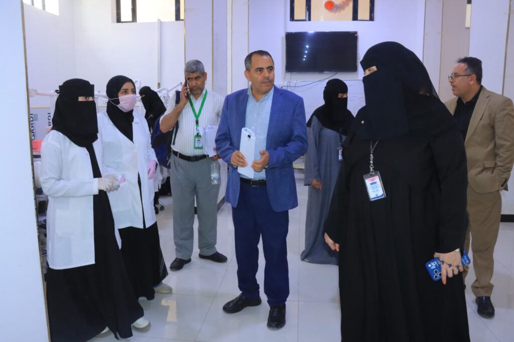 Group of professionals in a lab or hospital setting: two women in white coats and hijabs, a man in a blue suit, and others in scrubs or business attire, holding documents.