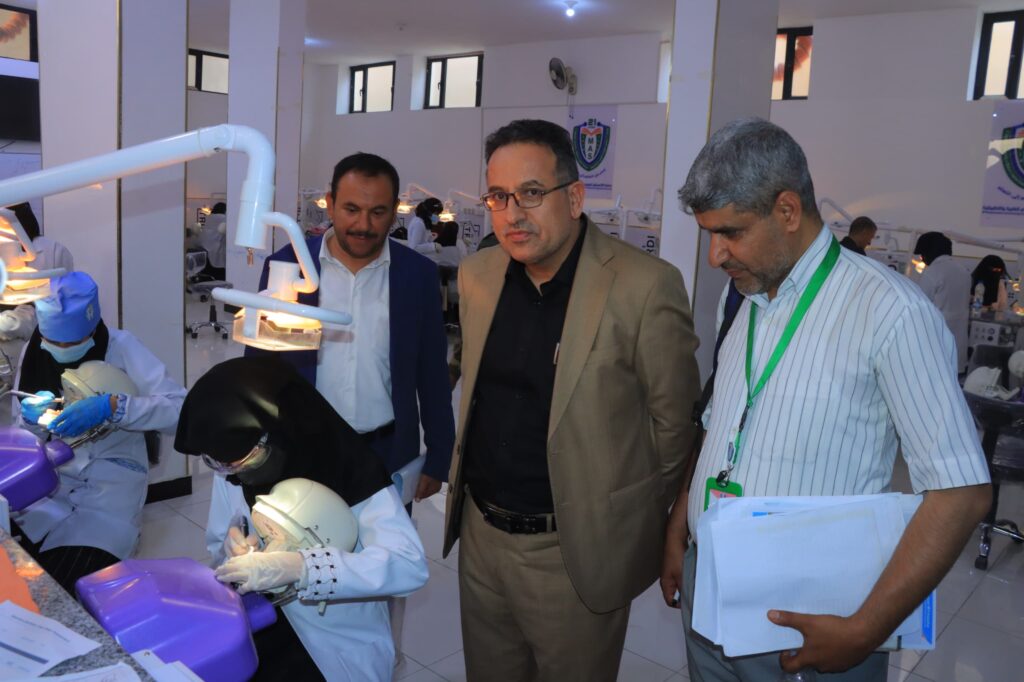Dental training session: staff and observers watch a student treat a patient at a dental chair in a bright clinic.