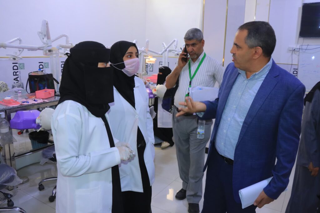 Two female dental professionals in hijabs and masks speak with a man in a blue suit in a busy dental clinic with chairs and equipment in view as they stand close together.