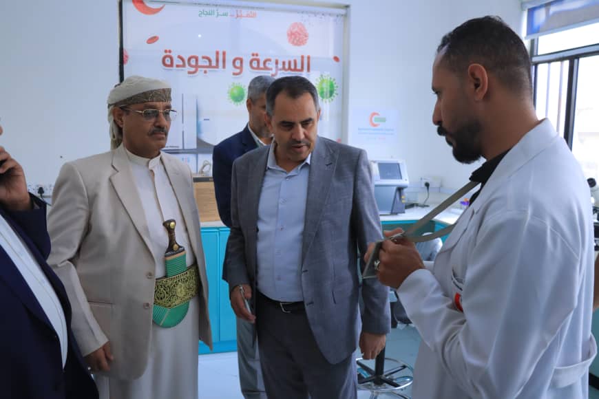 Group of men in a lab inspecting equipment; a technician in a white coat shows a device as others observe.