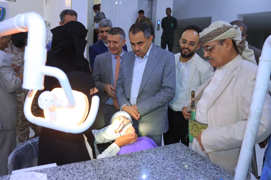 Dentist examines a patient under a dental light while several formally dressed men watch in a clinic corridor.