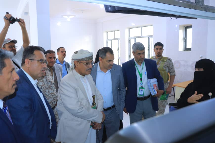 Officials and a woman in a niqab discuss a display in a bright, modern room with men in suits and uniforms nearby, during a tour.