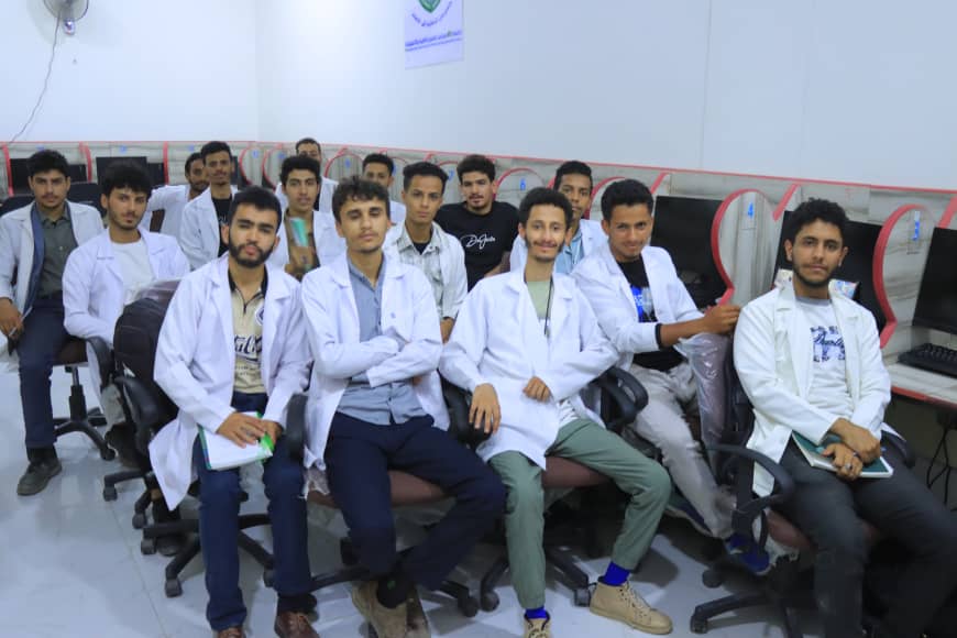 Group of young men in white lab coats posing in a computer lab with desks and PCs in the background.