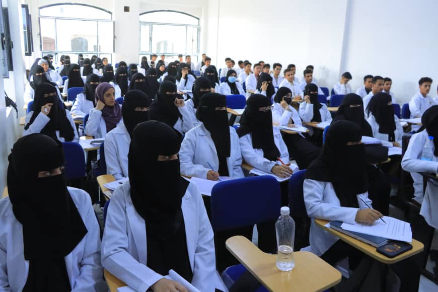 Crowded classroom of students in white coats; many women wearing black niqabs seated at desks taking notes in a lecture hall.