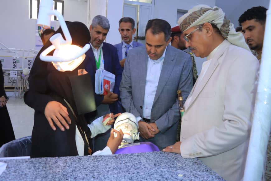 Female clinician wearing a hijab demonstrates a dental procedure on a skull model while a group of men watches nearby.