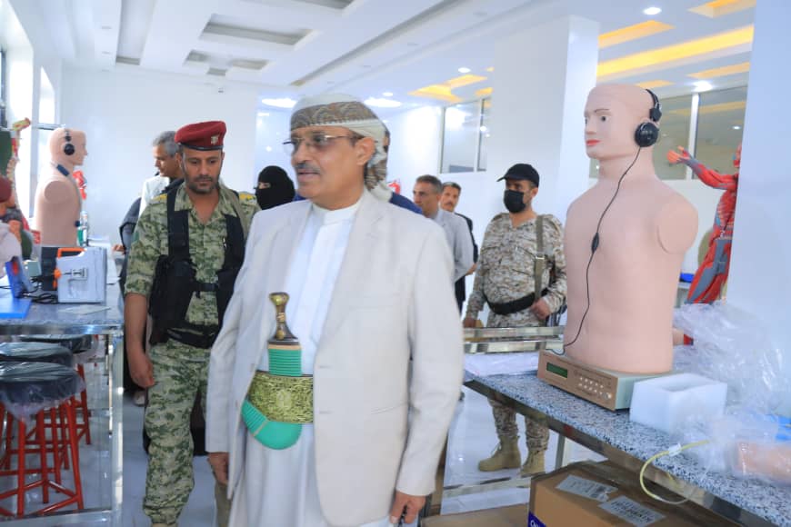 Group of men in a bright showroom; a man in white traditional attire with a ceremonial sword walks past while soldiers stand nearby, and a mannequin with headphones sits on a table in the background.