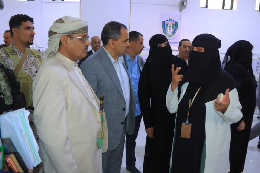 A woman in a black niqab and white lab coat explains something to a group of men in a bright, formal indoor setting.