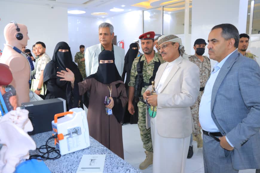 Group of adults, including a woman in a niqab, observing a medical training demo with a mannequin and equipment.
