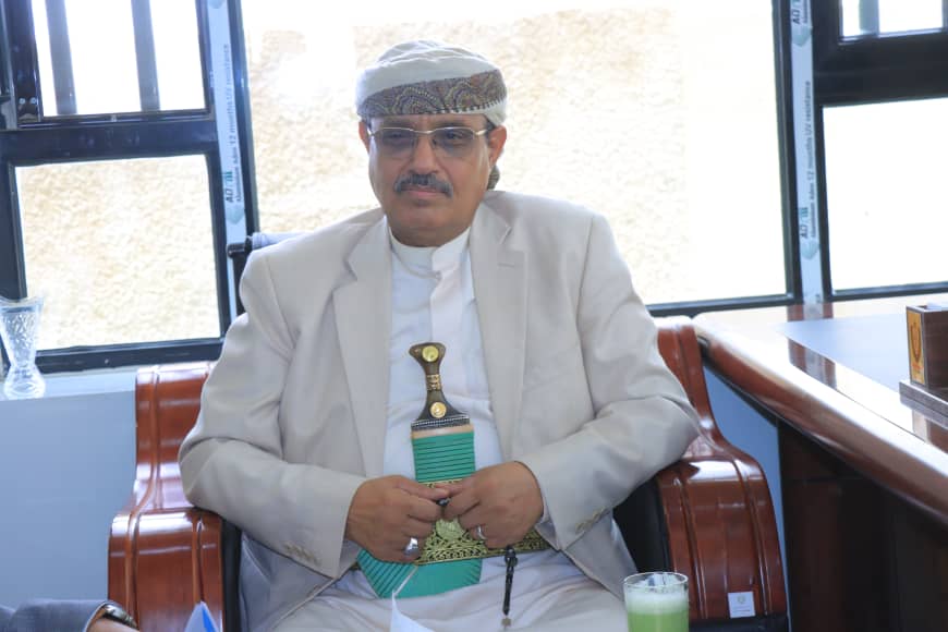 Man in white traditional attire and headscarf sits in an office chair with hands folded, a green drink on the table nearby, windows in the background.