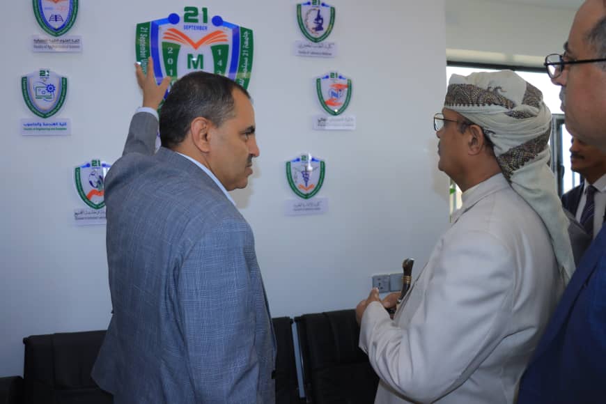 Two men in a formal setting face a wall of emblems as one places a hand on a crest.
