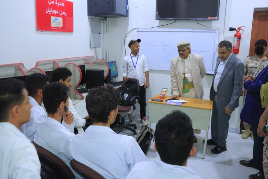 Students seated in white shirts watch a presenter in a beige suit at a small desk in front of a whiteboard.
