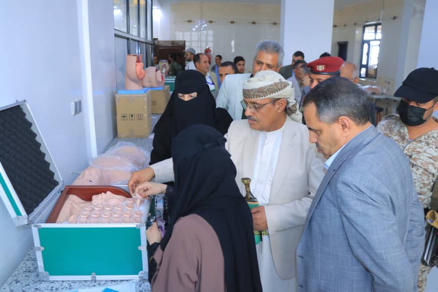 Group of people at a relief distribution center gathering around a green case with small cups or containers being prepared for distribution by a woman in a hijab and other officials nearby.