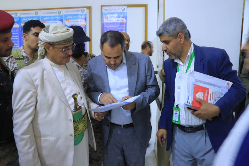 Three men gather around a document, with a man in a beige suit and traditional headwear pointing to the papers as the others review them nearby.