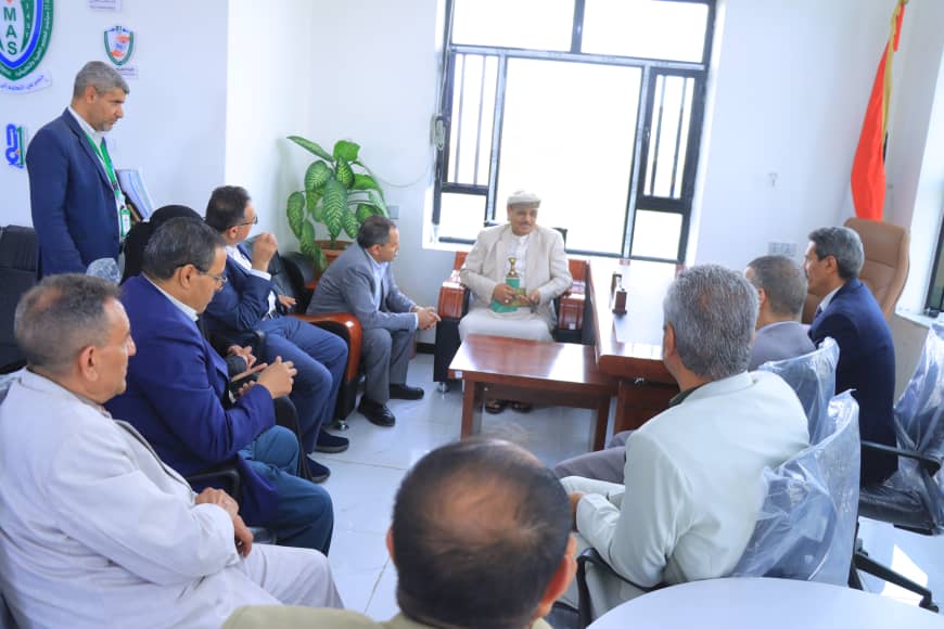 Elderly man in traditional white robes leads a meeting in a bright office while colleagues listen.