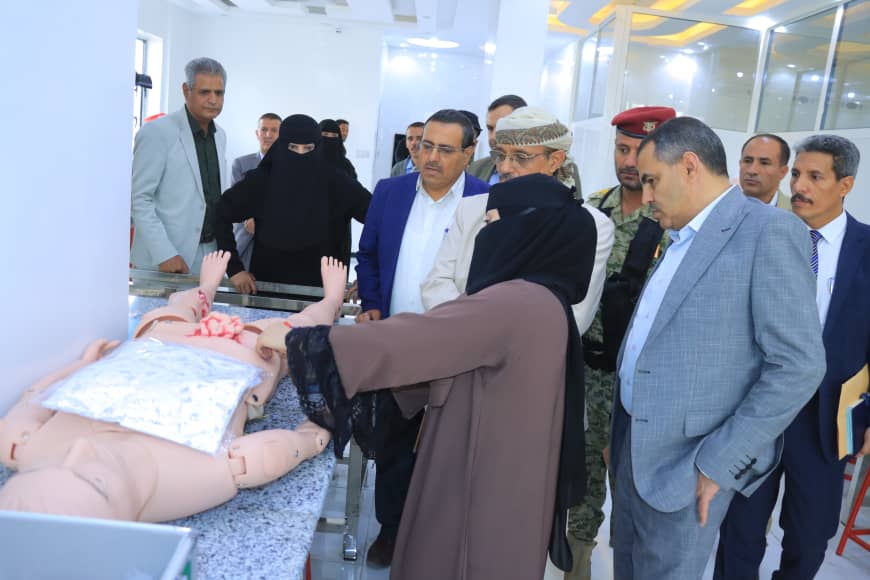 Group of officials and professionals observe a medical training mannequin laid on a table, as a woman in a hijab demonstrates a procedure in a clinical setting.