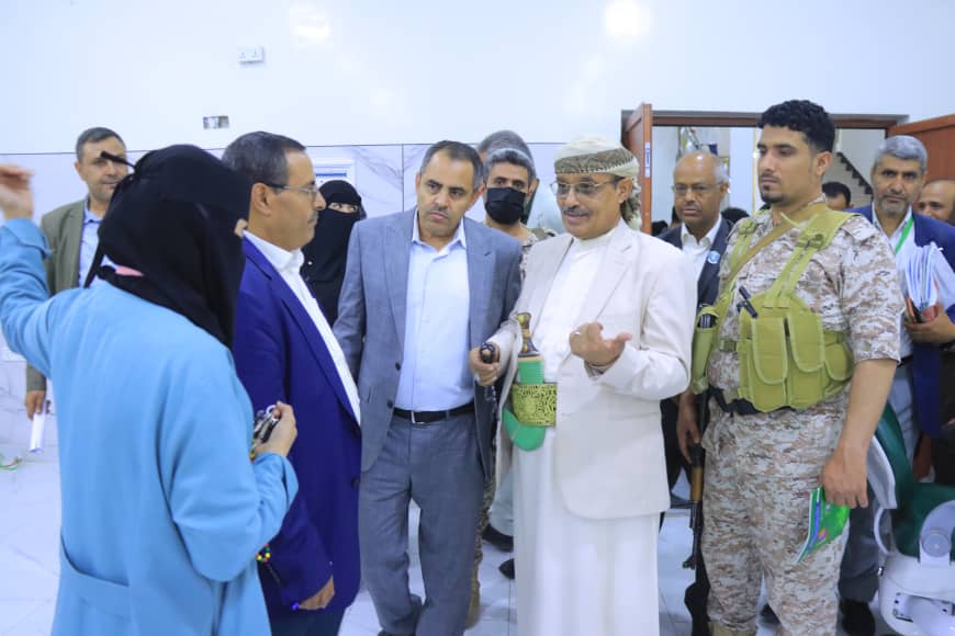 Group of officials and a soldier chat in a bright room; central figure in white traditional attire holds a green bottle and explains something.