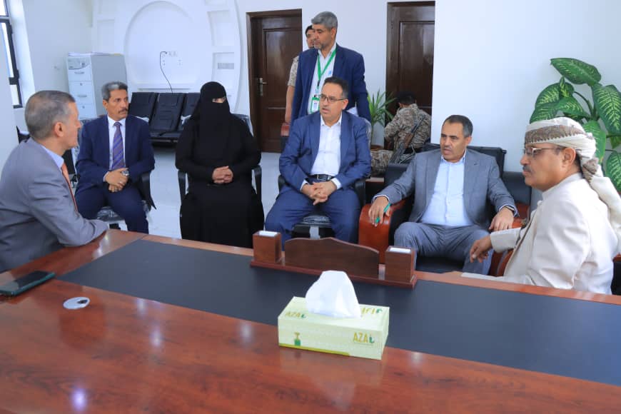 Seven people in a formal meeting around a large conference table; mix of suits and traditional attire in a bright office.