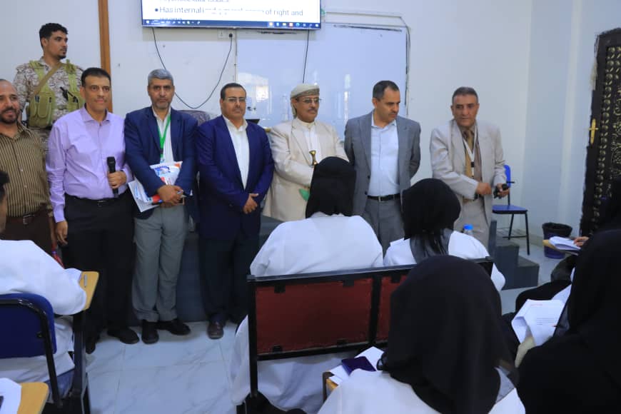 Group of men in business attire standing at the front of a classroom, facing seated female students in white coats who are taking notes on a marble-tiled room with a TV screen overhead.