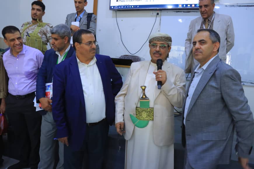 Group of men in a room posing for a ceremony; one man in traditional robe holds a microphone and a decorative trophy while others smile nearby.