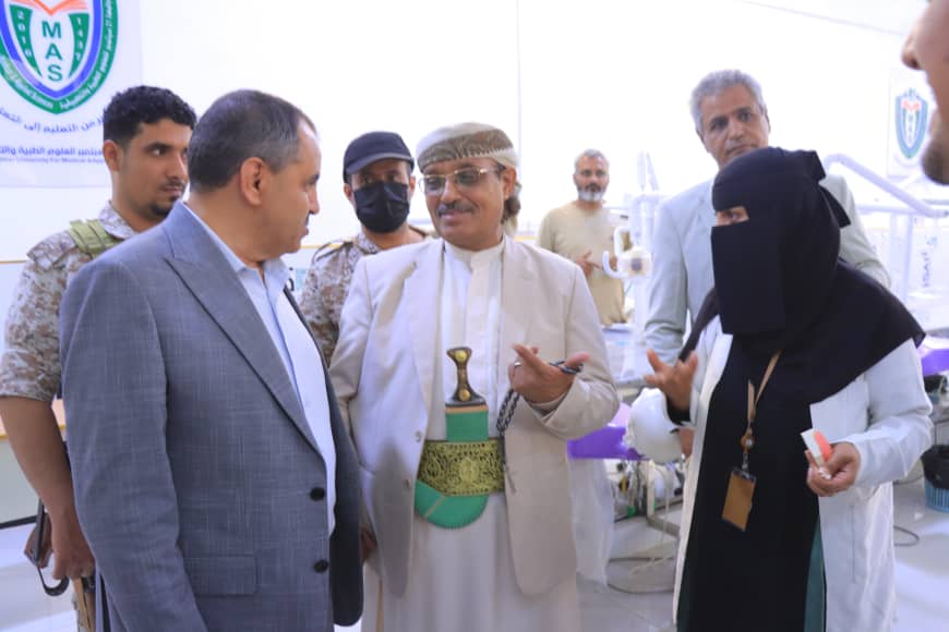 Group of officials and healthcare workers converse in a clinic setting; the man in the center wears traditional Yemeni dress and a ceremonial sword belt while talking to a man in a suit.",