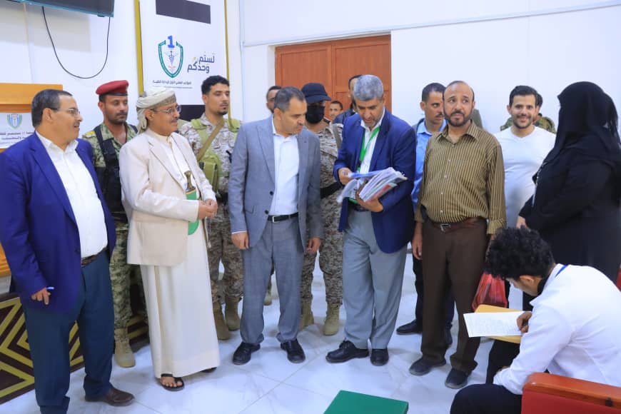 Group of officials and soldiers stand together in a hall; a man in beige traditional robes speaks with others while a woman in a black abaya stands on the right.