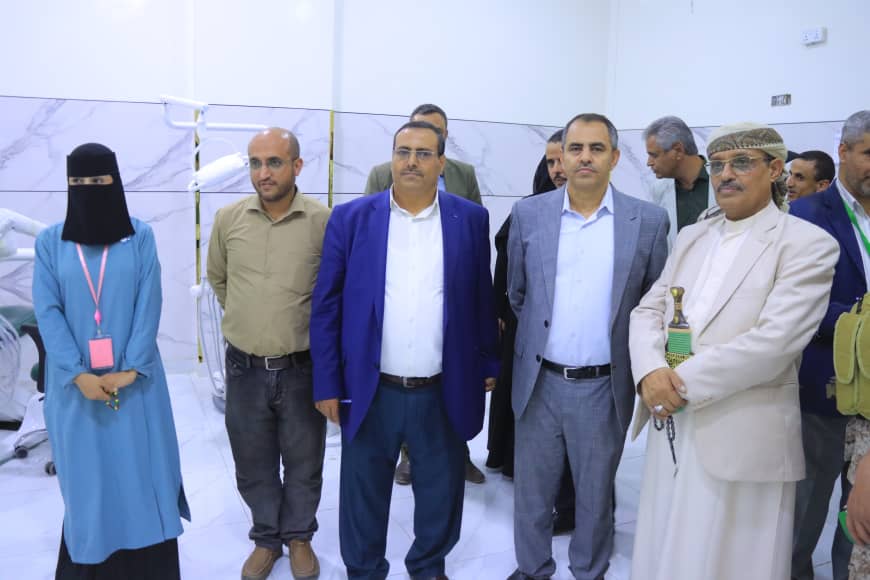 Group of seven people posing in a hospital-like lobby; a woman in a blue dress and black hijab stands at the left.