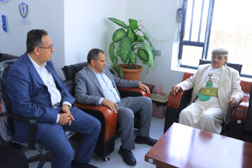 Three men sit in a bright office lounge having a discussion: two in suits and one in traditional attire, with a large potted plant nearby.