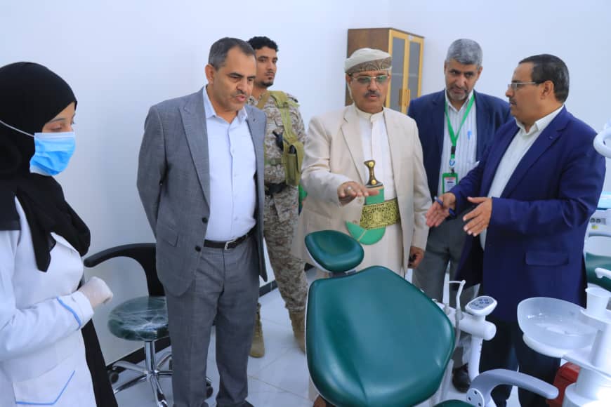 Group of men in suits and traditional attire stand around a green dental chair in a clinic, as a nurse in a mask looks on.
