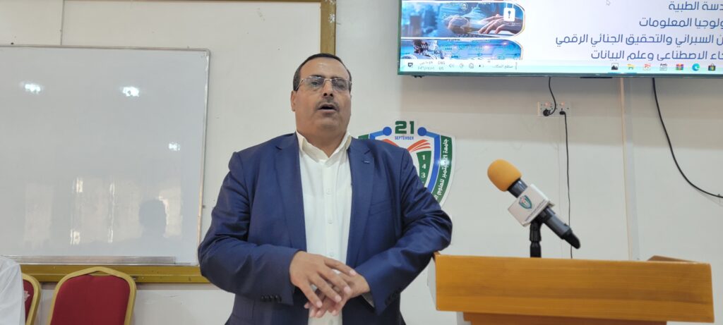 Man in a blue suit giving a speech at a wooden podium with a yellow microphone. Behind him is a whiteboard and a digital screen with Arabic text and a crest logo.