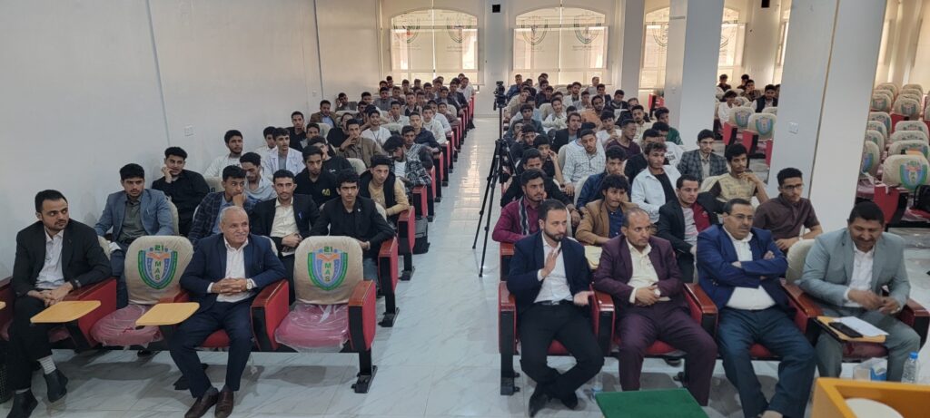 Audience of students and faculty seated in a lecture hall, a camera on a tripod in the center aisle.