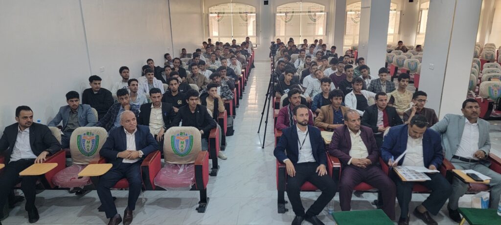 Large lecture hall full of attendees seated in rows, with a few men in suits in the front row and a camera on a tripod near the center.