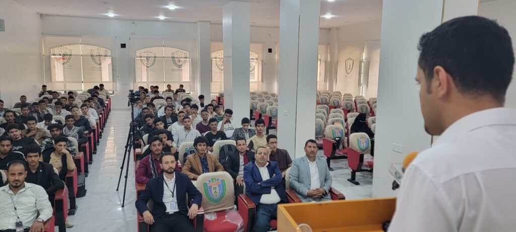 Speaker at a podium addressing a large audience of students in a bright lecture hall with red and beige seats.