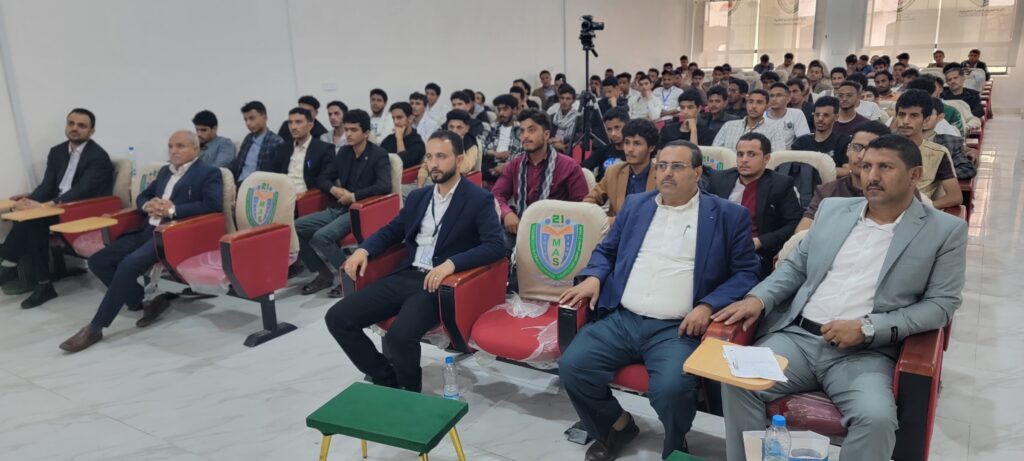 Audience at a conference: front row of men in suits, students behind, seated in a lecture hall with a tripod camera in the back left.