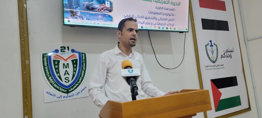 Man in a white shirt speaks at a wooden podium with a microphone, presentation screen and banners behind him at a university event.