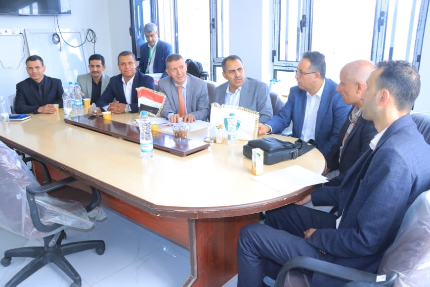 Group of men in suits seated around a curved conference table in a bright office, discussing with papers and drinks nearby. (informative meeting scene)