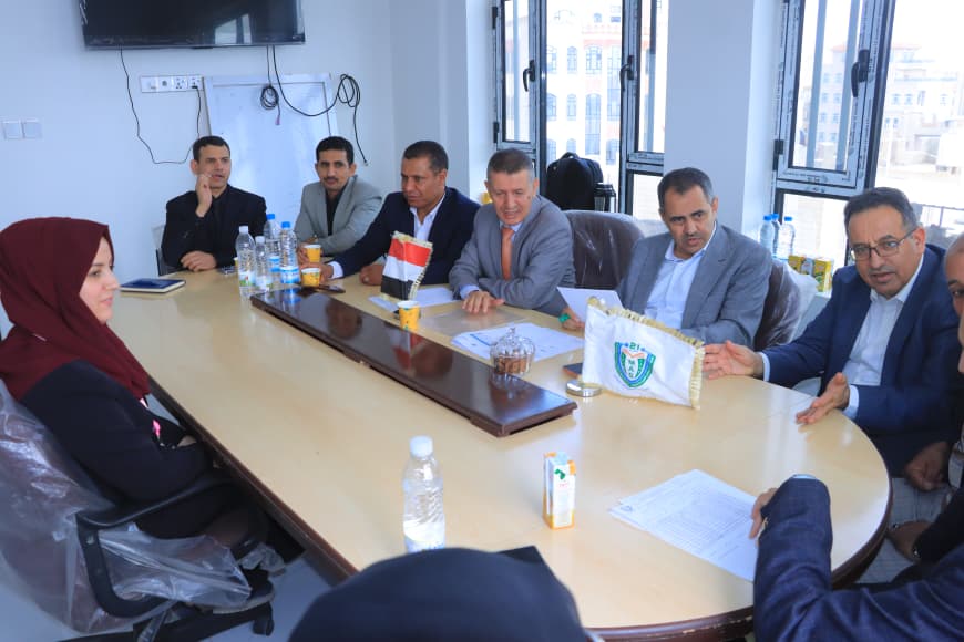 Group of professionals seated around a long conference table in a bright room, with flags and documents in the center as they discuss.