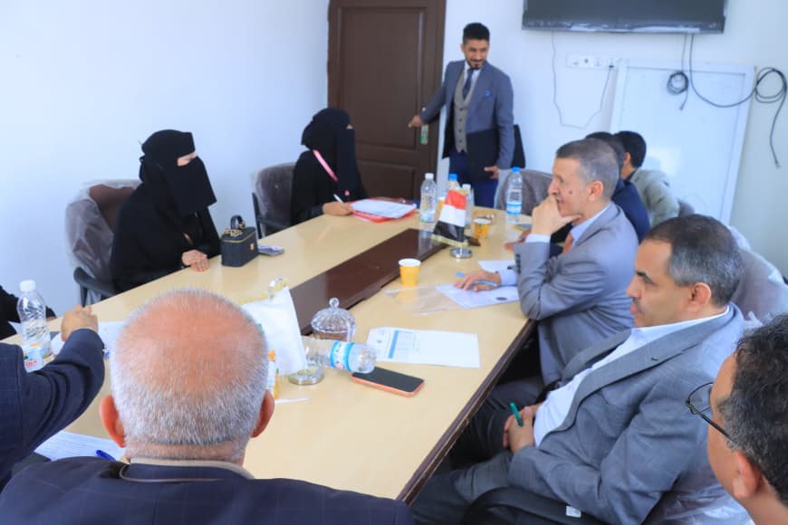 Formal meeting around a conference table; two women in black niqabs on the left, a man in a suit presenting near the door, and several men in suits with papers and water bottles.