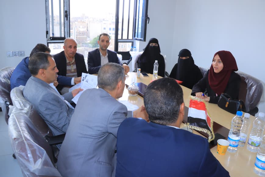 Group of professionals in a meeting around a table, with men in suits and women in headscarves, papers and water bottles on the table.