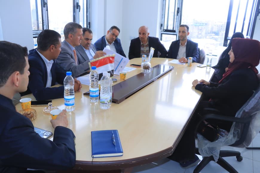 Group of professionals seated around a long conference table, reviewing documents with an Iraqi flag in the center and water bottles on the table.